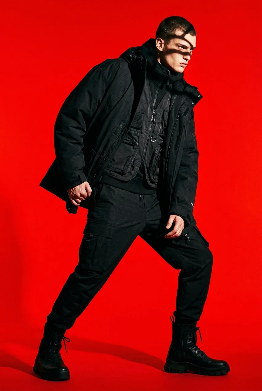 Model in head-to-toe black technical streetwear striding across a deep red studio backdrop