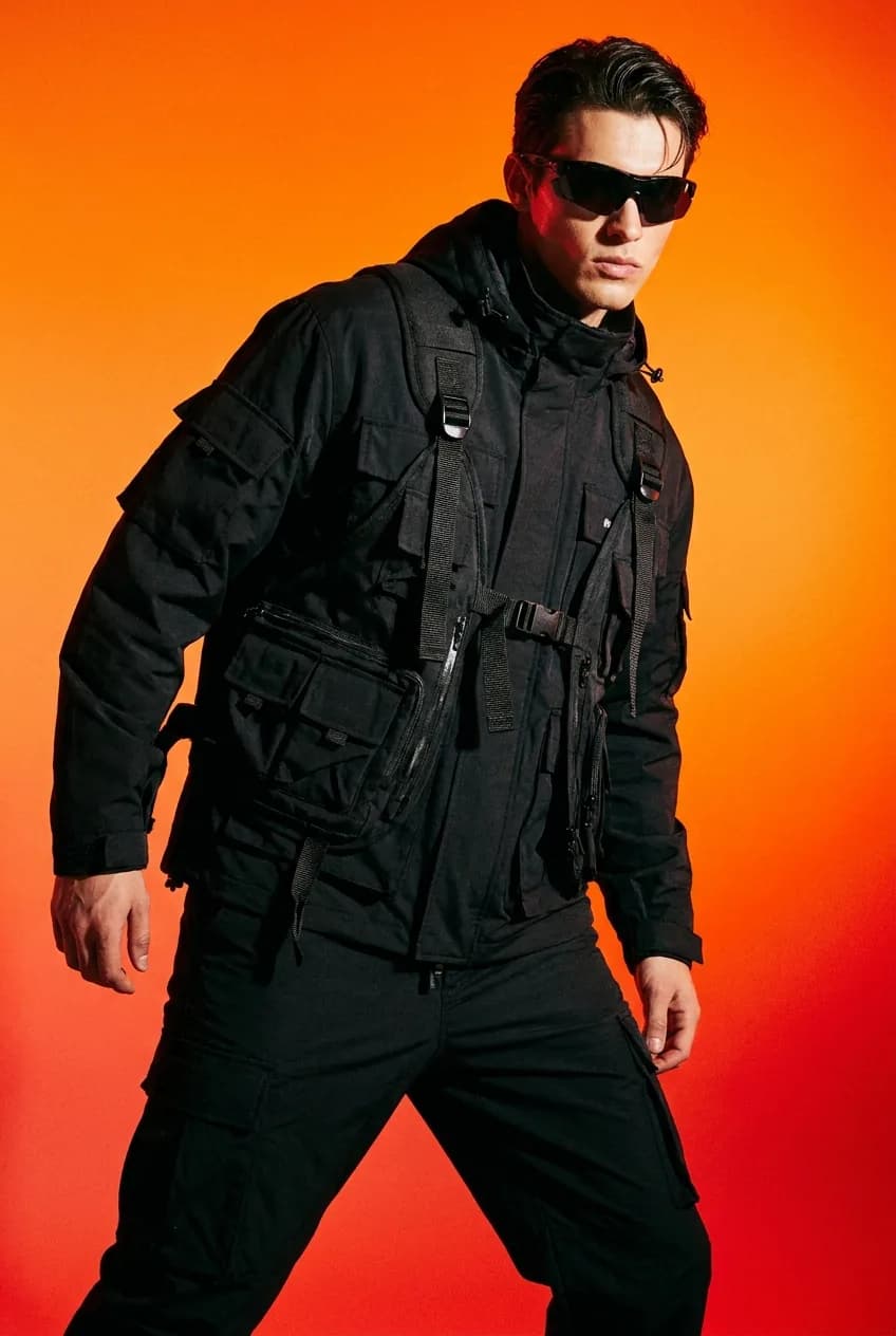 Model in a black utility rig and shell jacket against an orange-to-red gradient